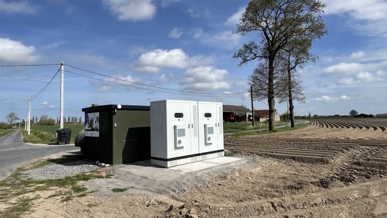Twee batterijkasten naast een transformatorhuisje op een erf in Vlaanderen, met een populier en een omgeploegde akker op de achtergrond