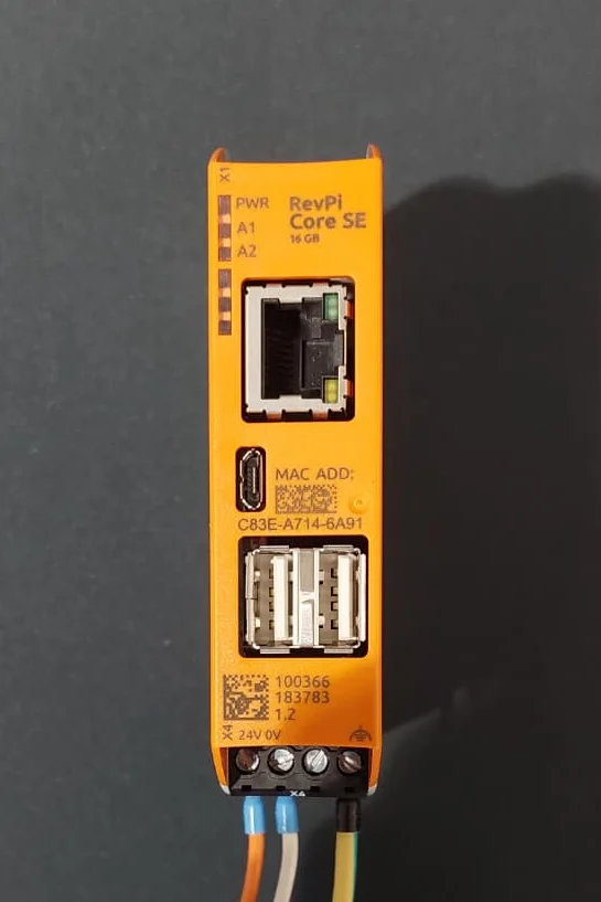 De EMS-controller, een RevPi Core SE, van voren met zijn statusleds, netwerkpoort, het label met het MAC-adres en de 24V-klem met draden in oranje, wit en geelgroen.