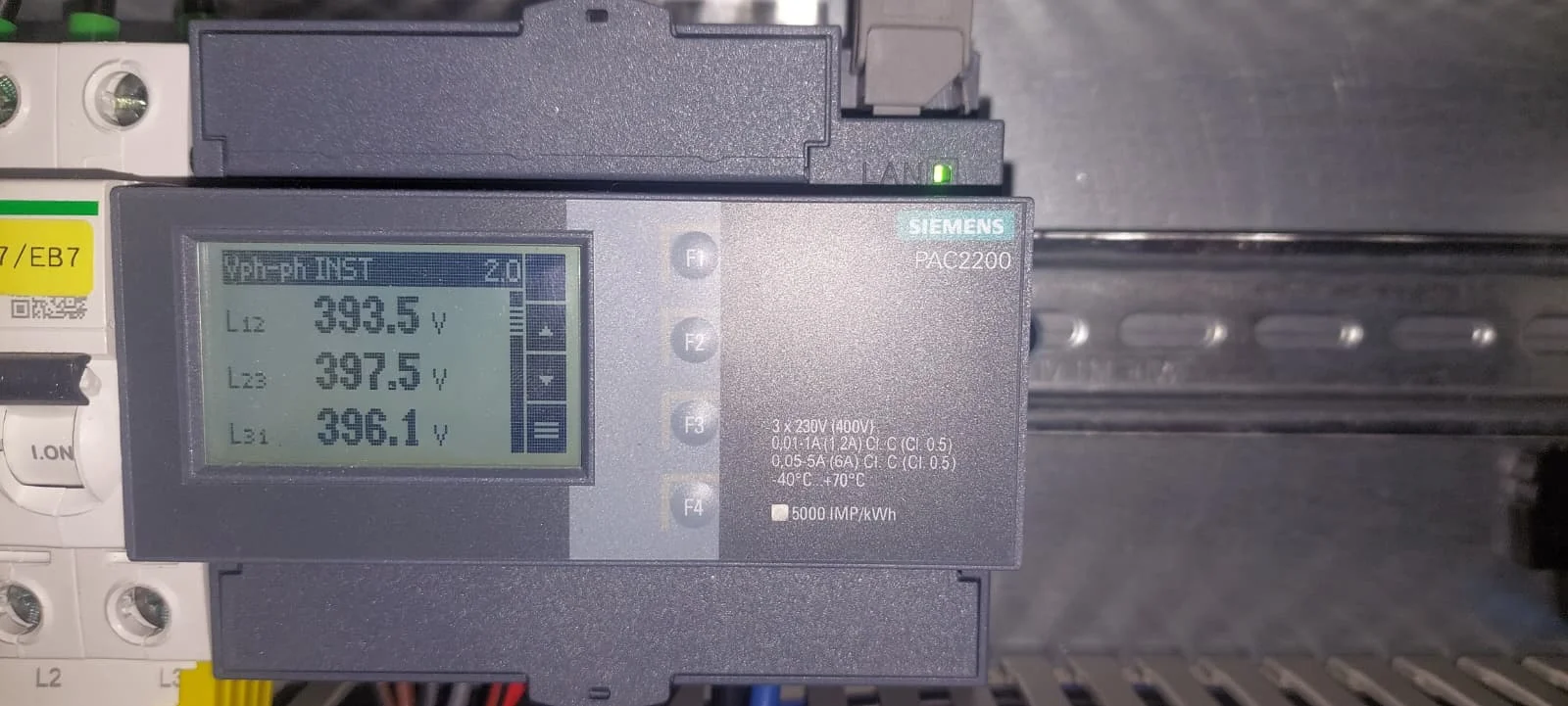 Een Siemens PAC2200 op een DIN-rail naast automaten, met op het display de momentane spanningen tussen de fasen L12, L23 en L31.